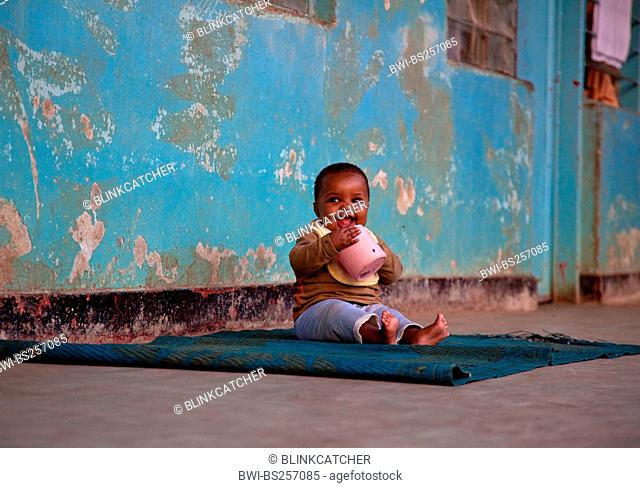 baby sitting on a carpet in front of the shabby wall of an orphanage sucking at a plastic pot, Burundi, Bujumbura Marie, Bujumbura
