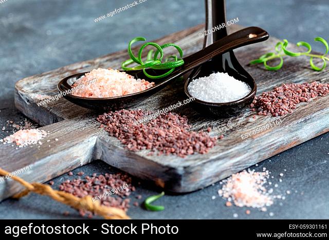 Spoons with white sea salt, pink Himalayan and black salt on wooden serving board, selective focus
