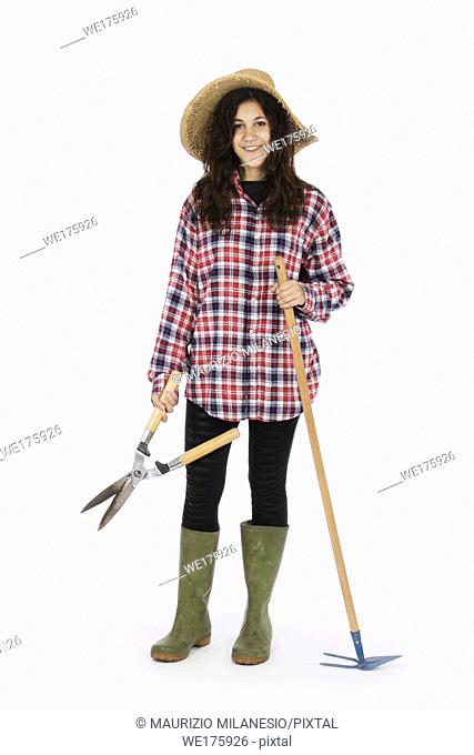 Young farmer with shears and hoe in hands, she is wearing a straw hat, checked shirt and green rubber boots