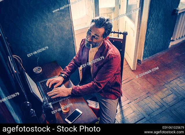 Busy Bearded Man With A Cigarette In His Mouth Types On The Laptop. Careless Middle-Aged Programmer Smokes And Drinks While Working From Home