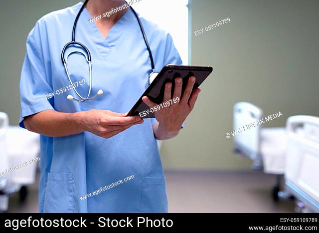 Midsection of caucasian female doctor in hospital wearing scrubs and stethoscope holding tablet