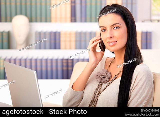 Portrait of attractive woman on mobile call sitting at home with laptop computer, smiling