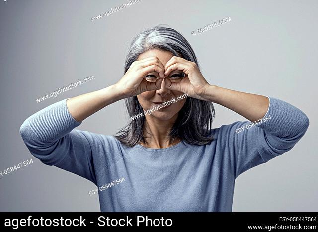 Beautiful Grey-Haired Woman with Dark Skin Squints Her Eyes While Looking Through Her Binoculars Like Hands. Attractive Asian in Light-Blue Sweater Posing in...