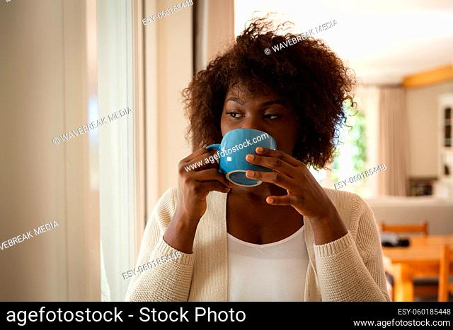 Thoughtful african american woman standing in dining room drinking cup of coffee