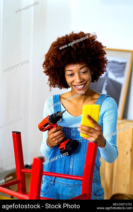 Smiling woman with electric screwdriver taking selfie through mobile phone at home
