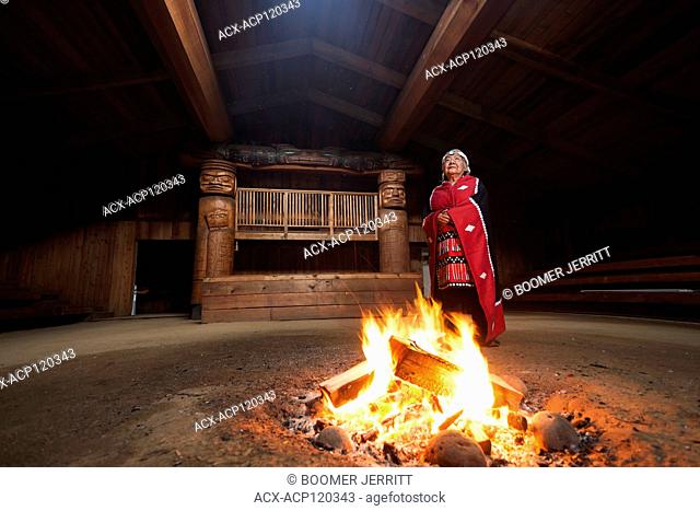 A first nations elder dressed in her traditional regalia stands beside a fire in the bighouse in the village of Oweekeno