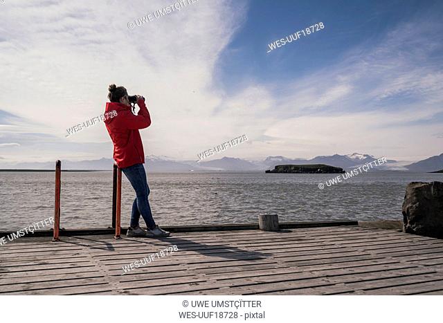 Young woman standig on a jetty, looking through binoculars, South East Iceland