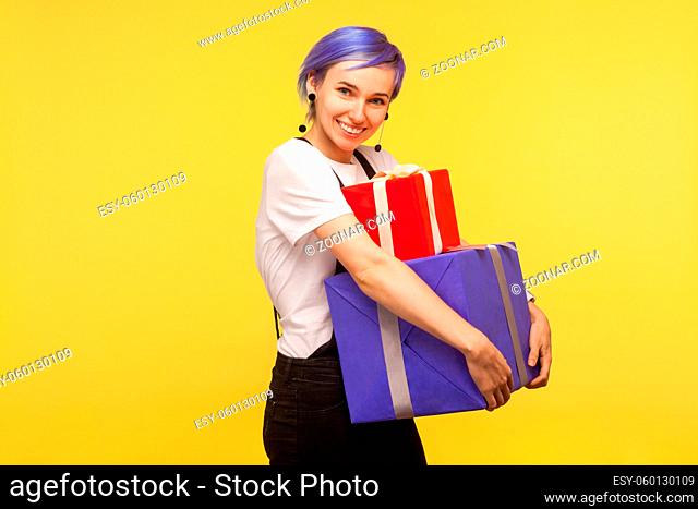 Portrait of glad positive hipster girl with violet short hair in overalls carrying gift boxes and smiling at camera, embracing holiday presents