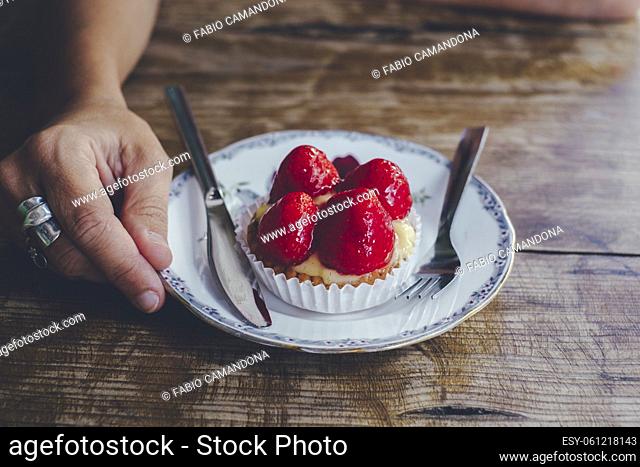 Close up of strawberry hand made tasty little cake and hand holding the dish on a wooden table. Concept of people and sugar natural fruits and food