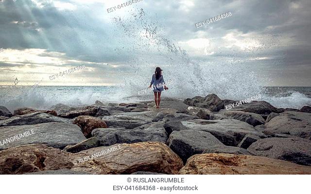 young woman standing on stones while big wave hitting shore