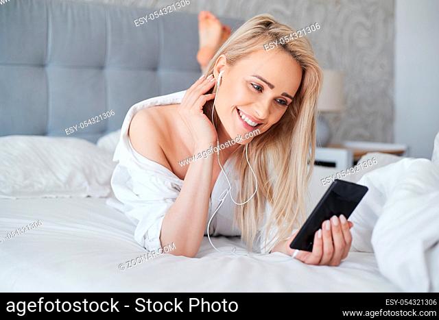 Beautiful middle aged, smiling blond woman lying in white bed and using a smartphone in her bedroom