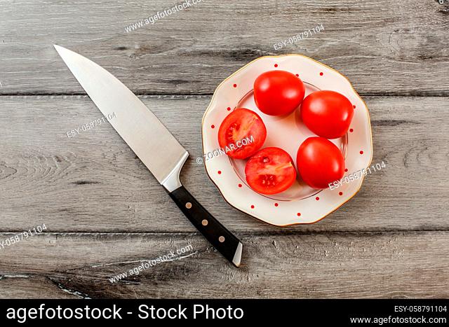 Tomatoes, one of them cut in half on white porcelain plate with small red dots, chef knife lying near. Tabletop view