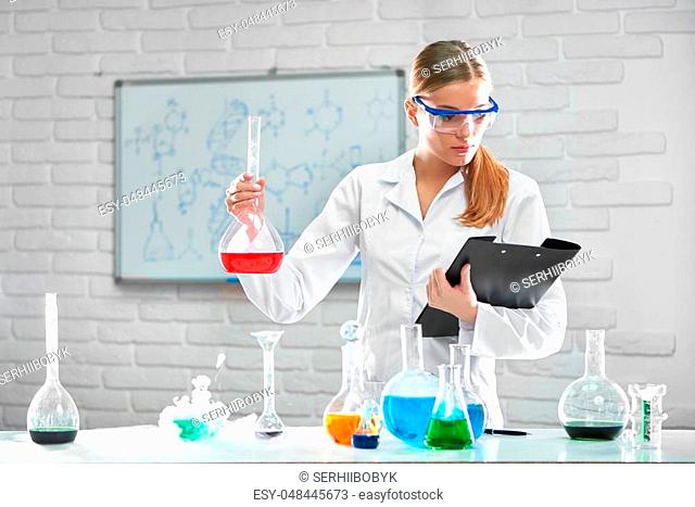 Shot of a beautiful young female researcher in a labcoat and protective eyewear reading her notes on a clipboard holding a flask with red chemical liquid...