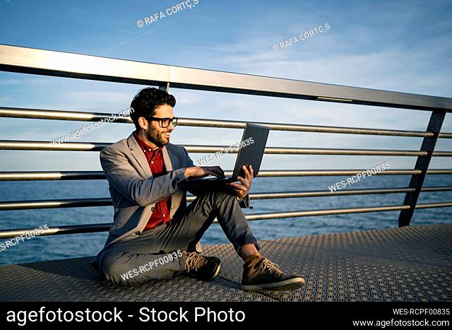 Smiling businessman using laptop while sitting on pier in front of railing