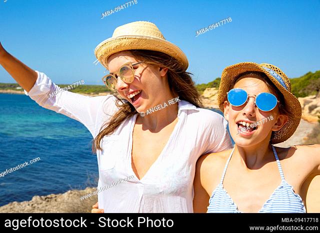 two girls in sunglasses and hats on the beach, laughing, waving