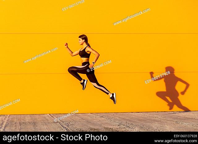 Young satisfied sporty beautiful woman with fit body jumping and running, hurry up against orange wall background. Gymnast jumping high with toothy smile