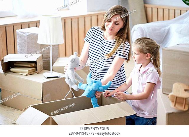 Pretty young mother and her cute little daughter having fun while sitting on floor of new apartment and unpacking moving boxes