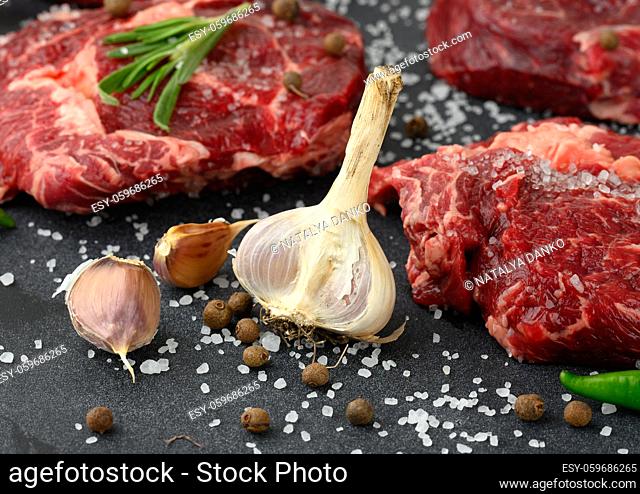 fresh garlic andraw piece of beef ribeye with rosemary, thyme on a black table, close up
