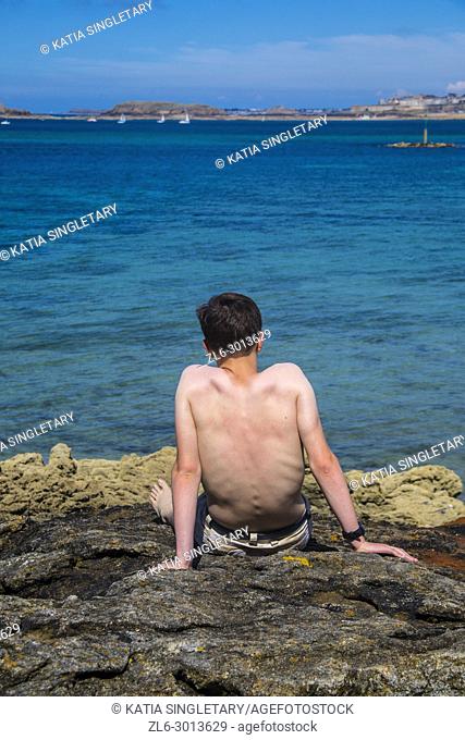 A caucasian male fair skin laying on rocks in front of the blue ocean. He is thinking, enjoying the peaceful view