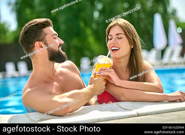 Lovely young woman sitting in the water and drinking the orange juice from the hands of the young man