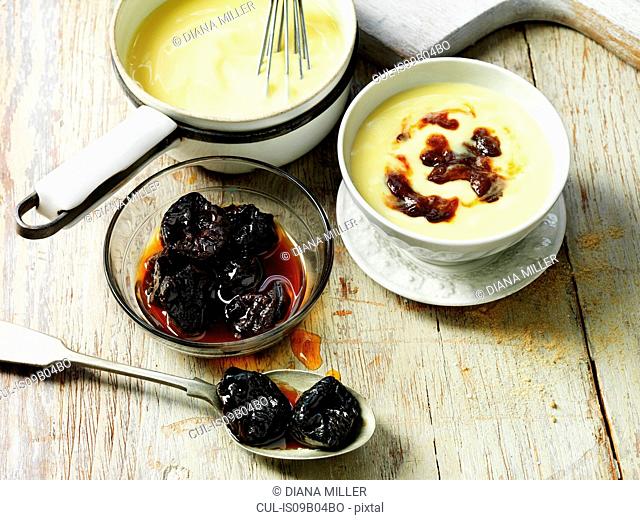 Prunes with custard in white bowl, prunes in spoon and glass bowl with syrup, custard in vintage pot, wooden table