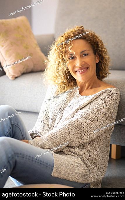 Portrait of happy young woman sitting on floor at home. Beautiful caucasian woman smiling sitting in living room. Pretty young woman with curly hair sitting in...