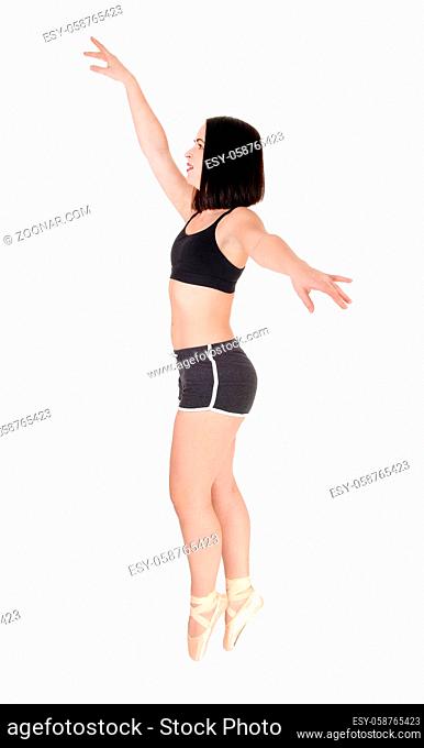 Slim young woman standing tiptoe in her ballet shoes and hands raised, in shorts and black top, isolated for white background