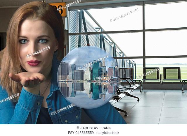 Sensual young woman is sending a kiss with a open palm at the airport terminal. The image of the Lower Manhattan skyline in New York City is in a transparent...