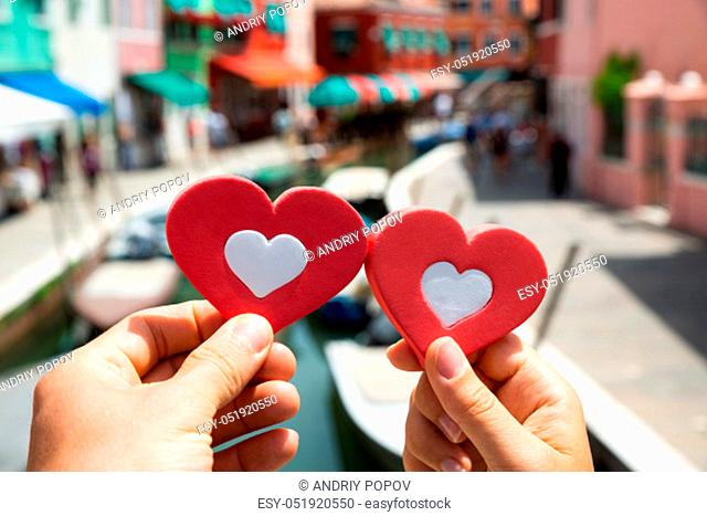 Close-up Of A Couple Hands Holding Red Hearts Shape Against Blurred Houses At Venice