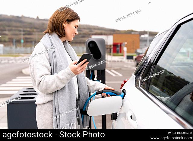 Woman with smart phone charging electric car at station