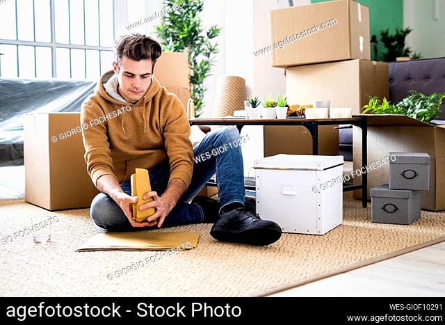 Young man wrapping drinking glass in brown paper while moving in new apartment