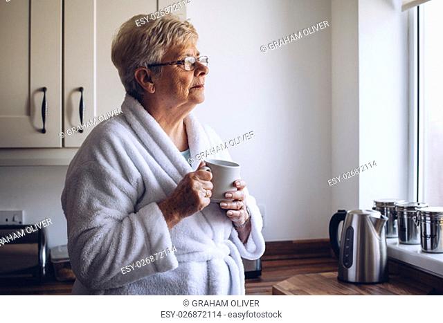 Senior woman looking out of her kitchen window with a cup of tea in hand