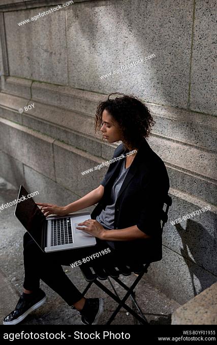 Afro female entrepreneur using laptop while sitting on chair in front of wall