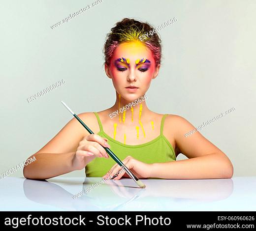 Portrait of young woman posing at the table with brush in hand. Unusual female art make-up with paint on brows, hair and around eyes