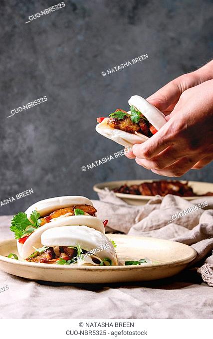 Man's hands hold asian sandwich steamed gua bao buns with pork belly, greens and vegetables served in ceramic plate on table with linen tablecloth
