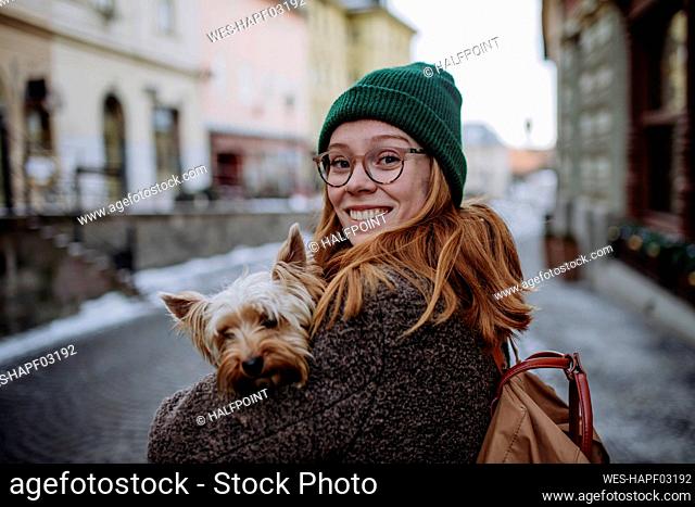 Happy redhead woman wearing knit hat carrying Yorkshire Terrier in city