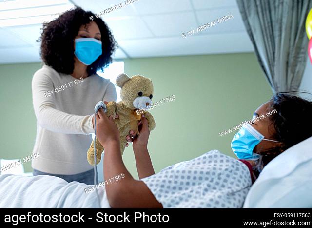Mixed race mother and sick daughter in face masks in hospital, playing with teddy bear
