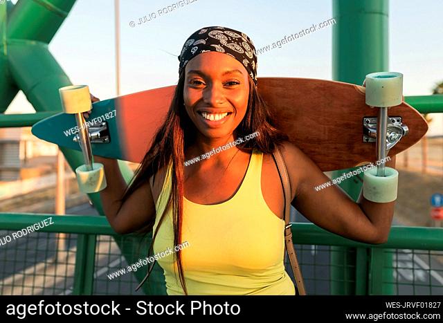 Smiling woman wearing headscarf carrying skateboard on bridge during sunset