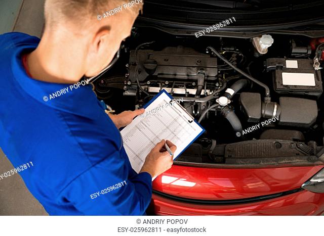 High Angle View Of A Mechanic Standing Near Car Writing On Clipboard In Garage