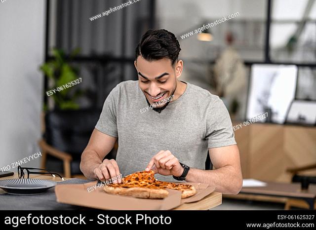 Snack. Joyful young adult man touching pizza in box sitting at table at home