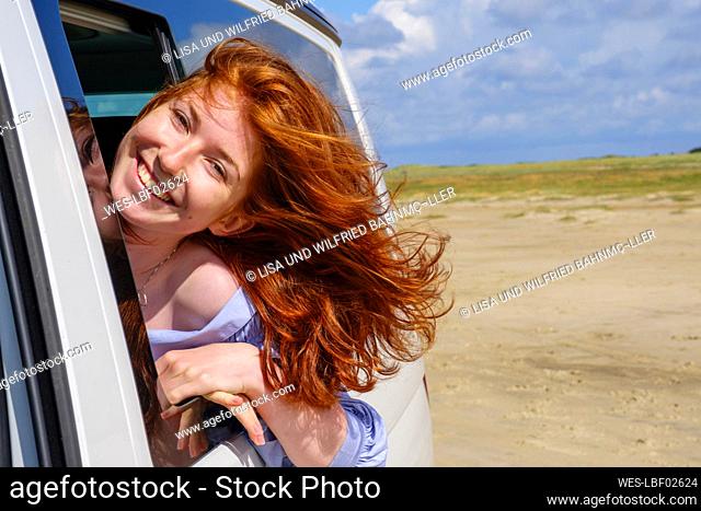 Portrait of carefree redhead teenage girl with head out of car window at beach against sky