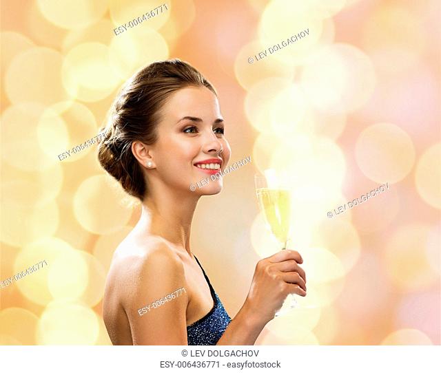 party, drinks, holidays, people and celebration concept - smiling woman in evening dress with glass of sparkling wine over beige lights background