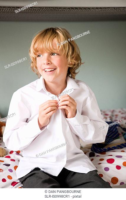 Young boy doing up the buttons of his school shirt while sitting on his bed