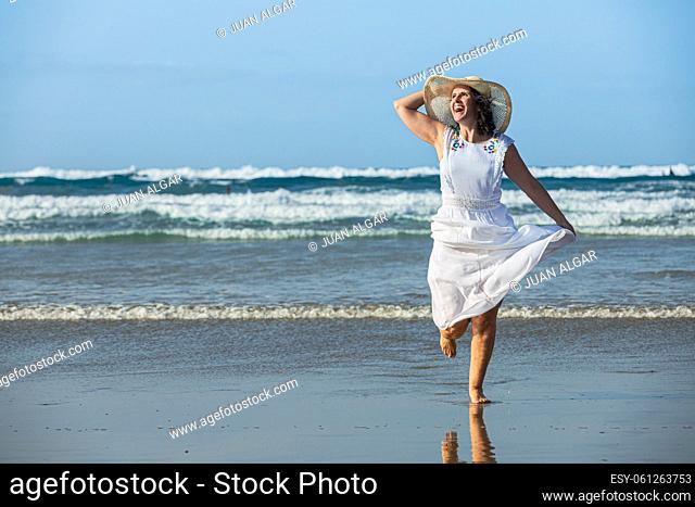 Optimistic female traveler in white dress laughing and looking away while running on wet sand against waving sea and blue sky