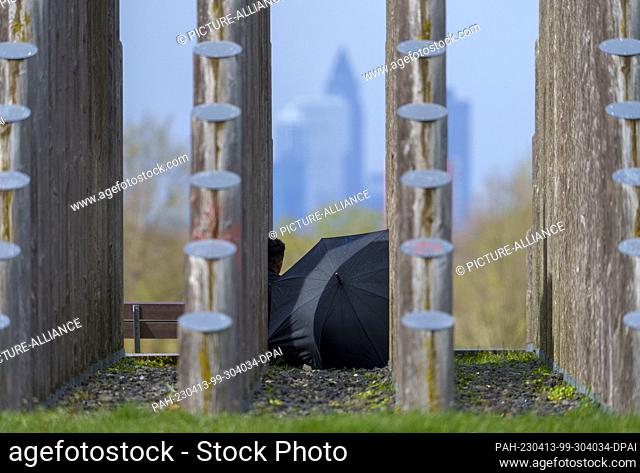 PRODUCTION - 13 April 2023, Hesse, Dreieich: A man sits under an umbrella between the tree trunks of the rod pyramid and looks at the skyline of Frankfurt am...