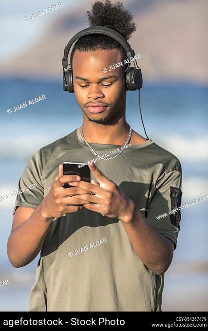 Handsome African American guy in modern headphones browsing smartphone and listening to music while standing on blurred background of sea