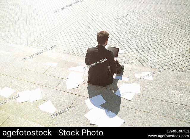 Toned picture of businessman sitting on stais and using laptop computer while many sheets of paper are all around. Business and freelance concepts