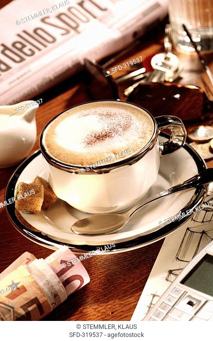 A cup of cappuccino amidst newspaper, keys and money