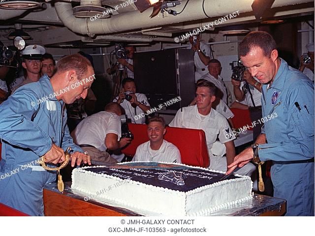 Astronauts Charles Conrad Jr. (left) and L. Gordon Cooper Jr. prepare to slice into the huge cake prepared for them by the cooks onboard the aircraft carrier...