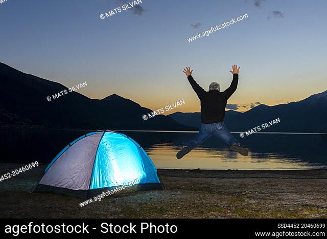 Man Jumping and an Illuminated Tent on Alpine Lake Maggiore with Mountain in Dusk in Ascona, Switzerland